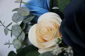 A close-up of a floral arrangement featuring creamy white and dark blue roses along with green eucalyptus leaves. The intricate layers of the rose petals are displayed beautifully, accompanied by the textured leaves and delicate small white flowers.