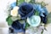 A vibrant bouquet of paper flowers in various shades of blue, teal, and white, surrounded by green foliage. The flowers are crafted with intricate detail, resembling roses and other blooms, with leaves adding a realistic touch.