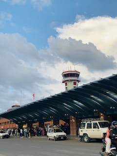 People gather outside an airport terminal building with a control tower in the background. Multiple vehicles, including cars and a motorcycle, are parked or moving near the terminal entrance. The sky is partly cloudy with blue and white cloud patterns.