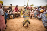 A vibrant outdoor scene with a group of people engaged in a celebration or dance. A woman is dancing energetically at the center while others gather around, clapping and watching. Several drummers provide music in the background, and people are dressed in colorful traditional clothing.
