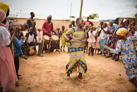 A vibrant outdoor scene with a group of people engaged in a celebration or dance. A woman is dancing energetically at the center while others gather around, clapping and watching. Several drummers provide music in the background, and people are dressed in colorful traditional clothing.