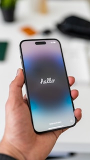 A hand holds a smartphone displaying a welcome screen with the word 'hello' prominently featured. The device has a sleek design with rounded edges and the background appears blurred, possibly indicating a depth effect. The screen has a gradient color scheme transitioning from dark at the top to lighter hues at the bottom.