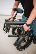 A person is holding a folded black electric bike with the brand logo 'G-Force' visible on the frame. The bike has thick tires and a sturdy build, suitable for portability. The setting appears to be outdoors, likely on a driveway.