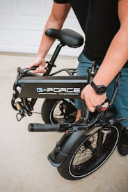 A person is holding a folded black electric bike with the brand logo 'G-Force' visible on the frame. The bike has thick tires and a sturdy build, suitable for portability. The setting appears to be outdoors, likely on a driveway.