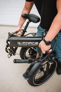 A person is holding a folded black electric bike with the brand logo 'G-Force' visible on the frame. The bike has thick tires and a sturdy build, suitable for portability. The setting appears to be outdoors, likely on a driveway.