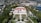 An elegant villa with a red-tiled roof is surrounded by manicured gardens featuring symmetrical paths and various plant life. The villa is positioned against a backdrop of lush greenery and a coastal cityscape, with a view of the calm blue sea.