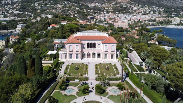 An elegant villa with a red-tiled roof is surrounded by manicured gardens featuring symmetrical paths and various plant life. The villa is positioned against a backdrop of lush greenery and a coastal cityscape, with a view of the calm blue sea.