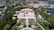 An elegant villa with a red-tiled roof is surrounded by manicured gardens featuring symmetrical paths and various plant life. The villa is positioned against a backdrop of lush greenery and a coastal cityscape, with a view of the calm blue sea.