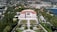 An elegant villa with a red-tiled roof is surrounded by manicured gardens featuring symmetrical paths and various plant life. The villa is positioned against a backdrop of lush greenery and a coastal cityscape, with a view of the calm blue sea.