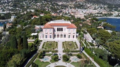 An elegant villa with a red-tiled roof is surrounded by manicured gardens featuring symmetrical paths and various plant life. The villa is positioned against a backdrop of lush greenery and a coastal cityscape, with a view of the calm blue sea.