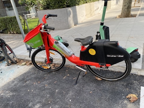 A brightly colored, shared electric bike branded with Lime and Uber logos is parked on a paved roadside. The bike has a red frame with green accents and a black chain guard. It features a front basket and is positioned on a bike stand next to a scooter. A concrete path with surrounding greenery is visible with some fallen brown leaves on the ground.