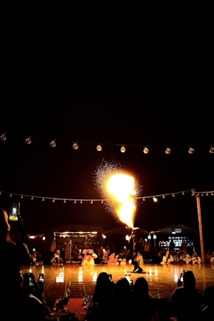 A performer is dramatically breathing fire in front of an audience, surrounded by string lights under a night sky. Several people are seated in a semi-circle, watching and capturing the moment with their phones. The scene is lively and filled with warm, glowing lights, highlighting the intensity and excitement of the performance.