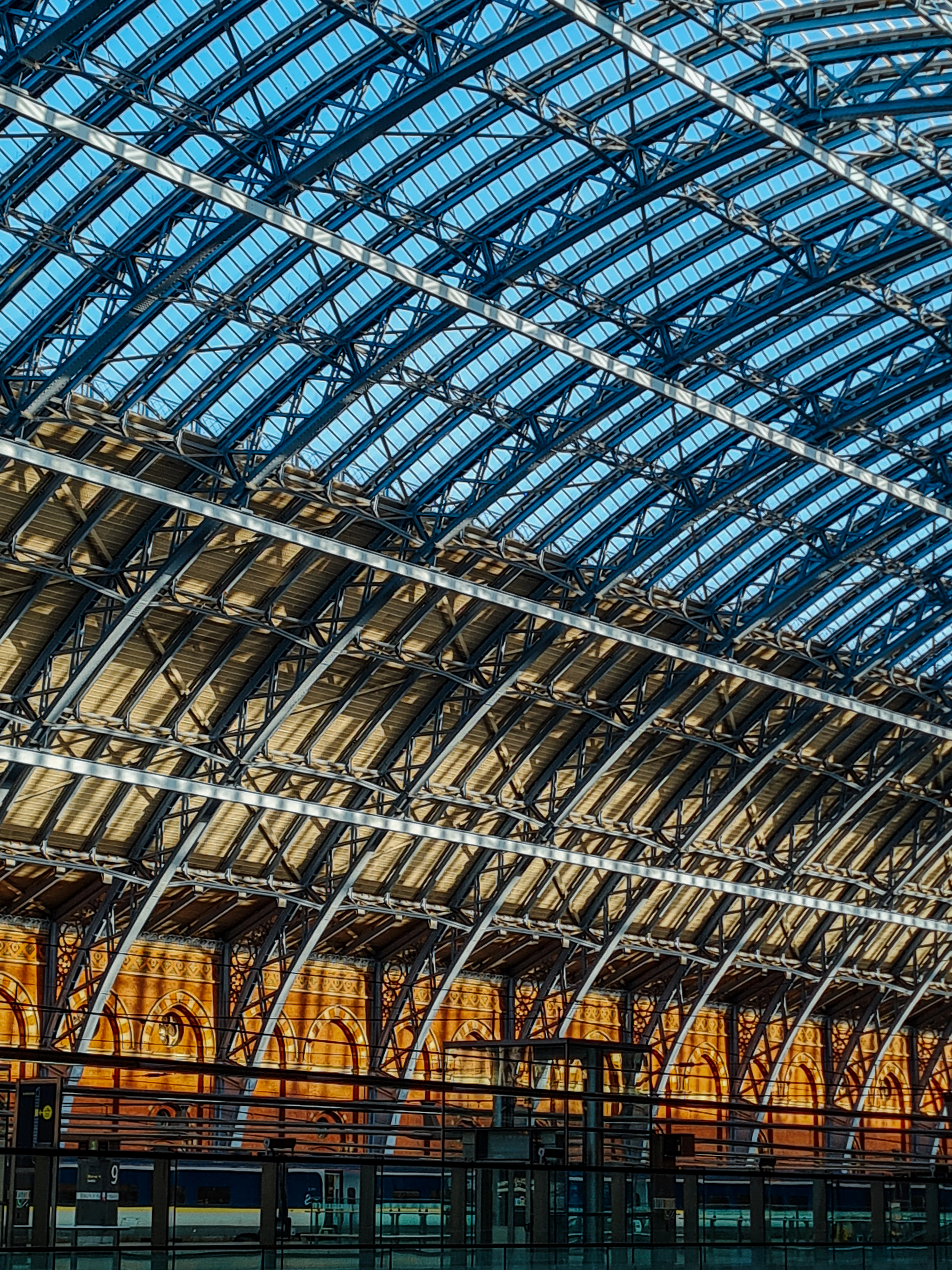 The Barlow train shed at St Pancras International seen from below: the great blue iron ribs of the arch springing across a glazed roof, sunlight on the ochre brick arcade at the far end and a Eurostar train waiting at platform nine