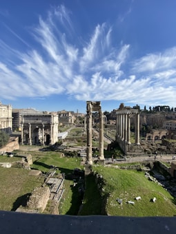 Ancient ruins dominate the landscape, with large columns and arches set against a vibrant blue sky. Sparse greenery is interspersed around the stone structures, indicating an archaeological site.