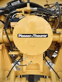 A detailed view of a yellow mechanical component with various hydraulic hoses and connectors. The central circular part has the label 'Plasser & Theurer'. The setup includes metallic bolts, pipes, and intricate wiring, indicating it is part of heavy machinery.