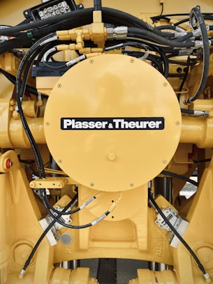 A detailed view of a yellow mechanical component with various hydraulic hoses and connectors. The central circular part has the label 'Plasser & Theurer'. The setup includes metallic bolts, pipes, and intricate wiring, indicating it is part of heavy machinery.
