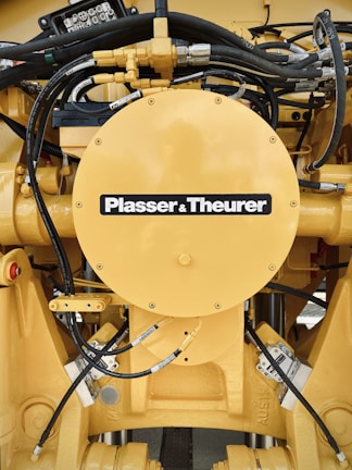 A detailed view of a yellow mechanical component with various hydraulic hoses and connectors. The central circular part has the label 'Plasser & Theurer'. The setup includes metallic bolts, pipes, and intricate wiring, indicating it is part of heavy machinery.