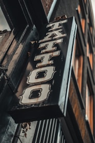 A vintage-style tattoo shop sign featuring bold, stylized lettering hangs at an angle against a building exterior. The sign has a distressed metal look with ornate designs around the letters.