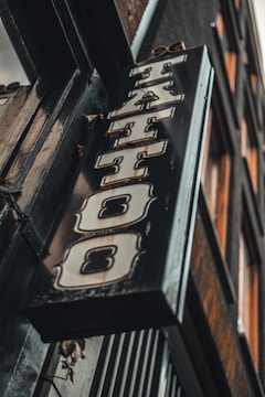 A vintage-style tattoo shop sign featuring bold, stylized lettering hangs at an angle against a building exterior. The sign has a distressed metal look with ornate designs around the letters.