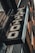 A vintage-style tattoo shop sign featuring bold, stylized lettering hangs at an angle against a building exterior. The sign has a distressed metal look with ornate designs around the letters.
