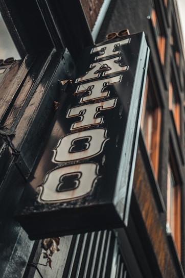 A vintage-style tattoo shop sign featuring bold, stylized lettering hangs at an angle against a building exterior. The sign has a distressed metal look with ornate designs around the letters.