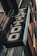 A vintage-style tattoo shop sign featuring bold, stylized lettering hangs at an angle against a building exterior. The sign has a distressed metal look with ornate designs around the letters.