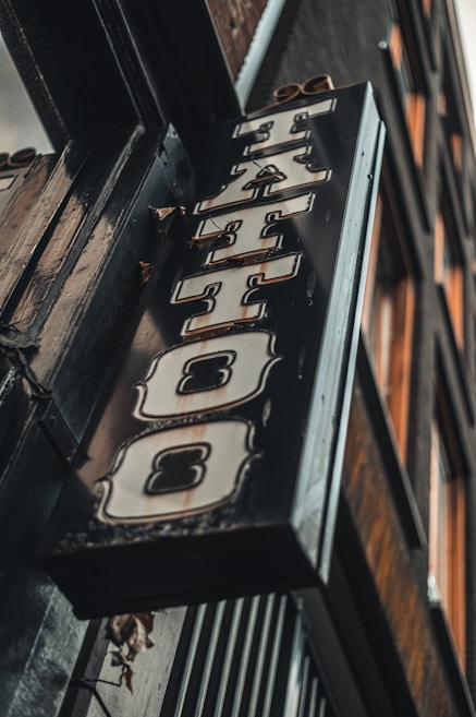 A vintage-style tattoo shop sign featuring bold, stylized lettering hangs at an angle against a building exterior. The sign has a distressed metal look with ornate designs around the letters.