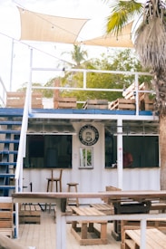 An open-air bar setup with wooden benches and tables made from pallets. The structure is white with a blue trim, featuring a small counter with a sign above it reading 'BARBA BLANCA Bar & Beerhouse'. There are two stools at the counter, and the bar is located under a canopy. Palms and greenery can be seen in the background, contributing to a tropical atmosphere.