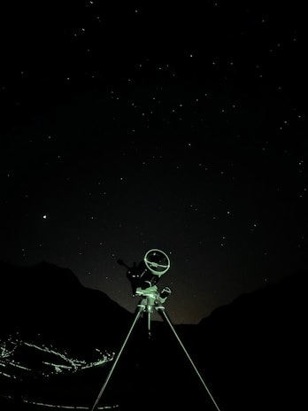 A telescope stands against a starry night sky, silhouetted by the faint outlines of mountainous terrain in the background. The foreground features a tripod supporting the telescope, illuminated subtly by ambient light.