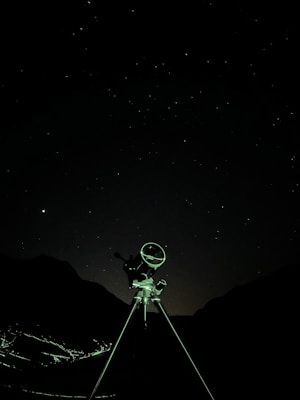 A telescope stands against a starry night sky, silhouetted by the faint outlines of mountainous terrain in the background. The foreground features a tripod supporting the telescope, illuminated subtly by ambient light.
