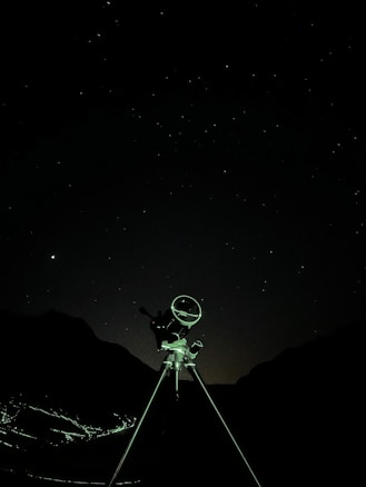 A telescope stands against a starry night sky, silhouetted by the faint outlines of mountainous terrain in the background. The foreground features a tripod supporting the telescope, illuminated subtly by ambient light.
