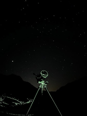 A telescope stands against a starry night sky, silhouetted by the faint outlines of mountainous terrain in the background. The foreground features a tripod supporting the telescope, illuminated subtly by ambient light.