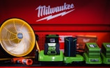 A display of various tools and equipment set against a red background with the Milwaukee logo. The items include a large yellow and black fan, several green battery chargers, and a red and black handheld tool. The setup is colorful and organized, suggesting a focus on industrial or workshop use.