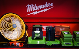 A display of various tools and equipment set against a red background with the Milwaukee logo. The items include a large yellow and black fan, several green battery chargers, and a red and black handheld tool. The setup is colorful and organized, suggesting a focus on industrial or workshop use.