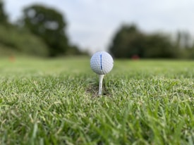 A close-up view of a golf ball balanced on a tee, situated on a patch of green grass. The background is blurred, revealing trees and possibly a course, suggesting a serene outdoor setting.