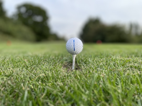 A close-up view of a golf ball balanced on a tee, situated on a patch of green grass. The background is blurred, revealing trees and possibly a course, suggesting a serene outdoor setting.