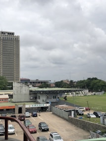 A large urban area featuring a tall building with a grid-like facade on the left. In the foreground, a parking lot is filled with several cars. Adjacent to the parking area is a structure with multiple signs, including one that reads 'TBSMB'. In the distance, a sports field and greenery are visible, bordered by trees and additional buildings. The sky is overcast with a blanket of grey clouds.