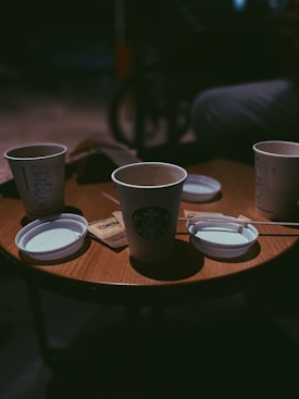 Several disposable coffee cups are placed on a wooden table in what appears to be a dimly lit cafe. The cups have lids nearby, and there are sugar packets and stirrers scattered on the table surface. The lighting creates a cozy and intimate atmosphere.