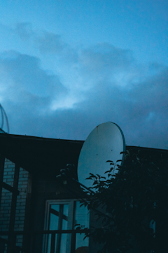 A satellite dish is prominently featured against a backdrop of a cloudy sky at dusk. The silhouette of a building and some foliage is visible in the foreground.