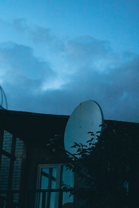 A satellite dish is prominently featured against a backdrop of a cloudy sky at dusk. The silhouette of a building and some foliage is visible in the foreground.