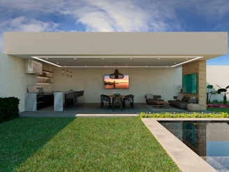 A modern outdoor living space featuring a covered patio with a ceiling fan and integrated lighting. To the left, there is an outdoor kitchen equipped with a grill, countertops, and bar stools. The center has a dining table with chairs, and on the right is a lounge area with a sofa and coffee table. The patio overlooks a pool and lush green lawn. There are decorative plants and a blue sky with some clouds.
