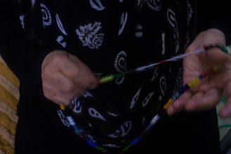 An individual is holding and possibly stringing a colorful beaded necklace or bracelet. They are wearing a black garment with a white pattern that includes various designs such as paisley and floral. The background is partially blurred, consisting of what appears to be a wooden surface on one side.
