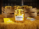 A jar of premium German blossom honey with a yellow and white label is prominently displayed in the foreground. The jar features a golden lid and is surrounded by a rustic wooden crate filled with similar jars. The scene is set on a surface covered with straw, and a card with the brand name 'Bienen Konig' is placed on the straw.