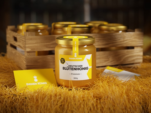 A jar of premium German blossom honey with a yellow and white label is prominently displayed in the foreground. The jar features a golden lid and is surrounded by a rustic wooden crate filled with similar jars. The scene is set on a surface covered with straw, and a card with the brand name 'Bienen Konig' is placed on the straw.