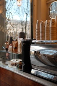 A cozy café setting featuring a shiny espresso machine with a stack of paper cups on it. Nearby, there are various pastries displayed in a glass case, and a window shows an outdoor view with trees. The environment suggests a warm, inviting atmosphere.