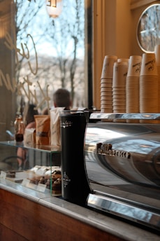 A cozy café setting featuring a shiny espresso machine with a stack of paper cups on it. Nearby, there are various pastries displayed in a glass case, and a window shows an outdoor view with trees. The environment suggests a warm, inviting atmosphere.