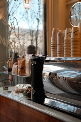 A cozy caf&eacute; setting featuring a shiny espresso machine with a stack of paper cups on it. Nearby, there are various pastries displayed in a glass case, and a window shows an outdoor view with trees. The environment suggests a warm, inviting atmosphere.