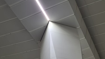 An architectural interior with sharp geometric lines formed by the ceiling and wall angles. Light panels illuminate the space, creating a modern and minimalist aesthetic.
