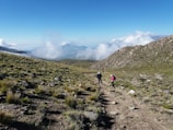 A rugged mountain landscape with hikers walking along a dirt path. Green vegetation covers the rocky terrain, and clouds hang low over distant peaks in the clear blue sky.