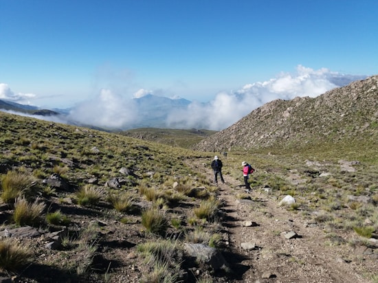A rugged mountain landscape with hikers walking along a dirt path. Green vegetation covers the rocky terrain, and clouds hang low over distant peaks in the clear blue sky.